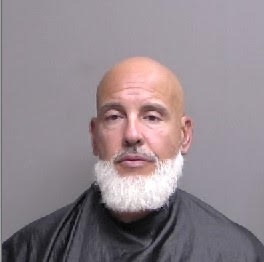 A bald man with a prominent white beard is shown against a plain background He is wearing a dark cape like garment The image is a straightforward headshot, and his expression is neutral