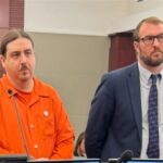 A man in an orange jumpsuit and another man in a suit stand in a courtroom A police officer and a woman stand in the background They are in front of a wooden panel with microphones