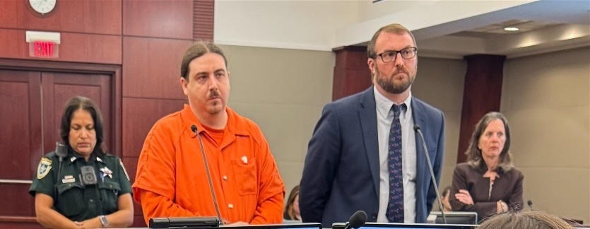 A man in an orange jumpsuit and another man in a suit stand in a courtroom A police officer and a woman stand in the background They are in front of a wooden panel with microphones