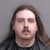 A man with long brown hair, a mustache, and a light beard, wearing a black shirt, standing against a plain gray background