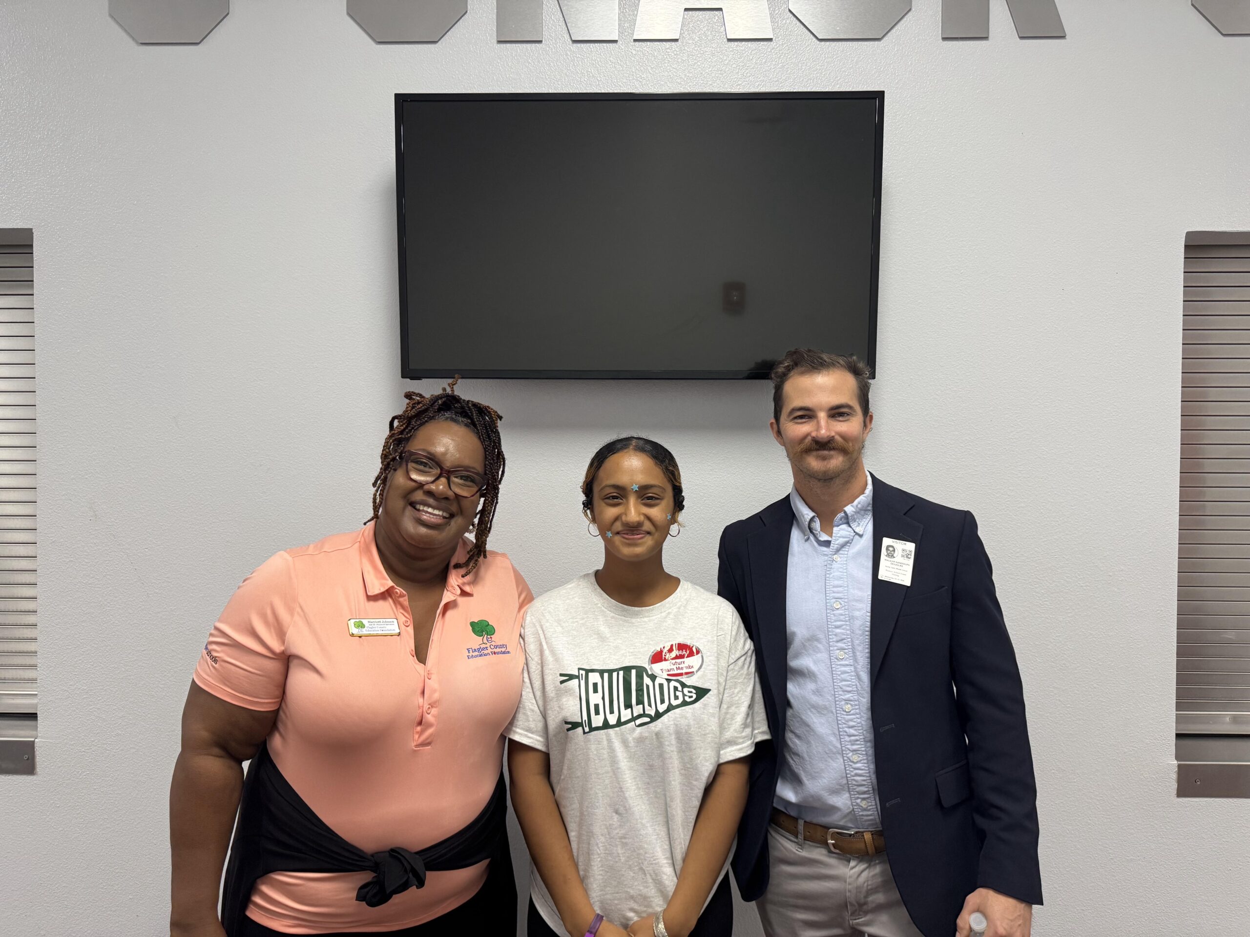 Three people stand smiling in front of a wall with the word "SNACK" partially visible above them The person on the left wears a peach blouse, the middle person wears a white t shirt with "BULLDOGS" text, and the person on the right is in a suit