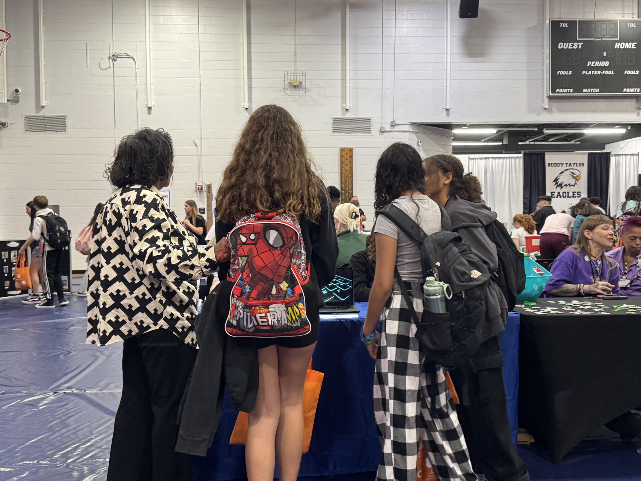 A group of people stand at a table in a gymnasium filled with others One person wears a Spider Man backpack The floor is covered with blue mats, and there are booths in the background A scoreboard hangs on the wall