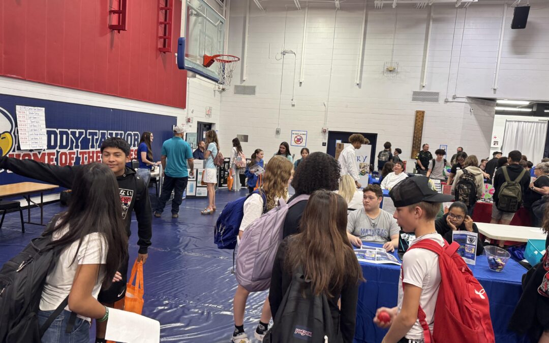 Students gather in a gymnasium for a school event They engage with representatives at various booths, some holding tote bags and pamphlets The gym is decorated with banners and a basketball hoop is visible