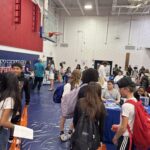 Students gather in a gymnasium for a school event They engage with representatives at various booths, some holding tote bags and pamphlets The gym is decorated with banners and a basketball hoop is visible