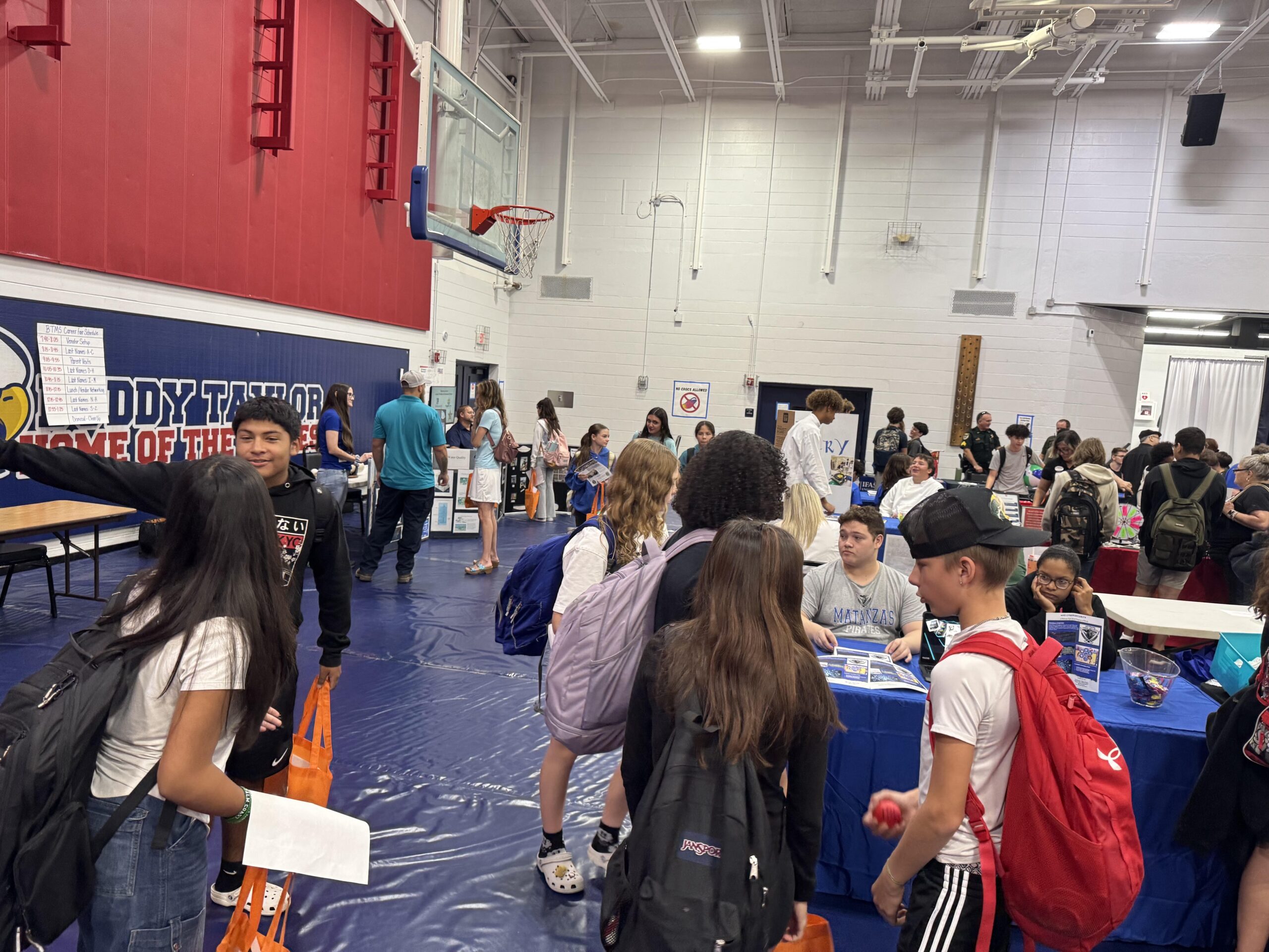 Students gather in a gymnasium for a school event They engage with representatives at various booths, some holding tote bags and pamphlets The gym is decorated with banners and a basketball hoop is visible