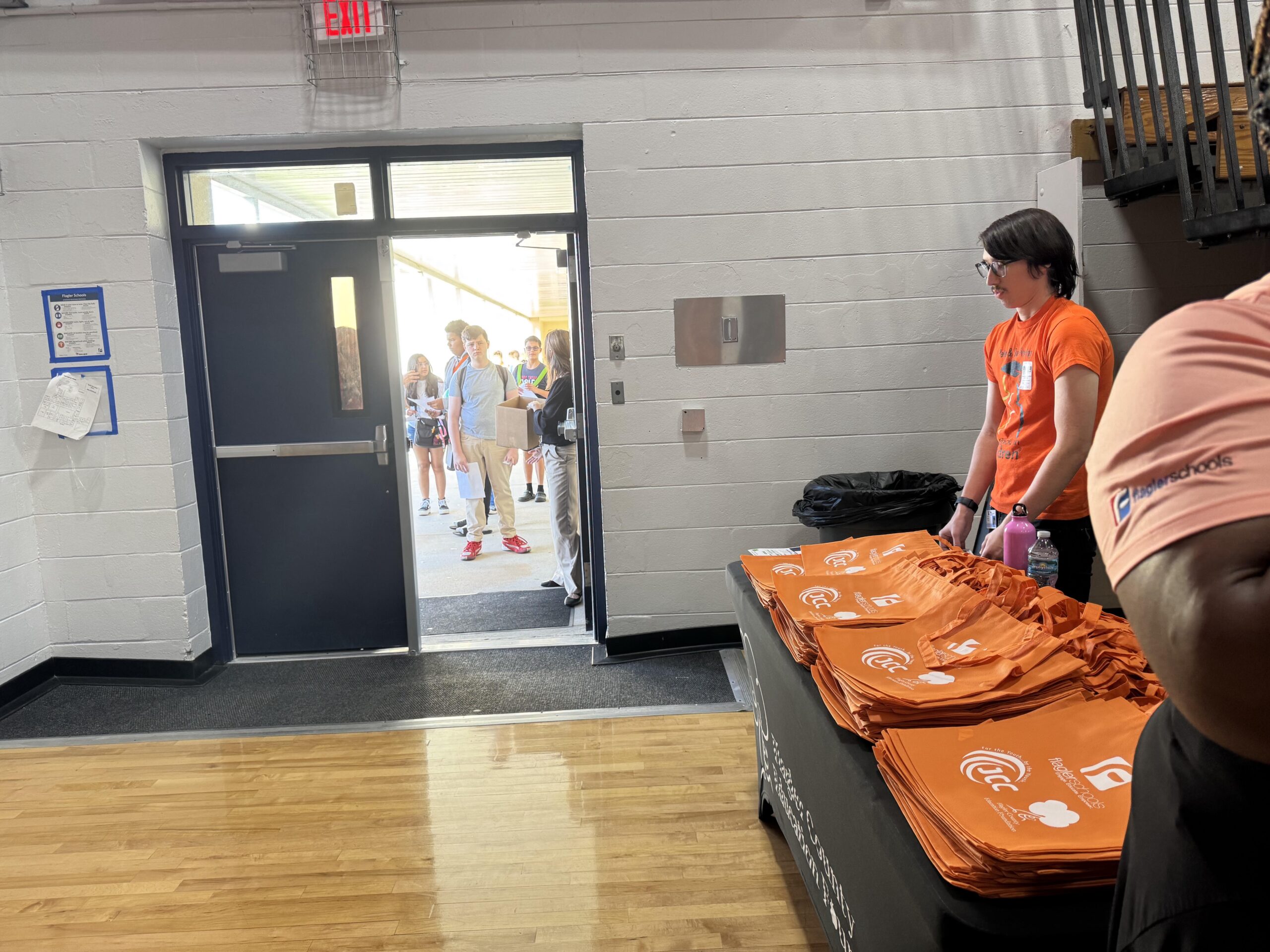 A person stands next to a table covered with orange drawstring bags in a hallway A group of people is visible outside the open door The room has wooden flooring and white brick walls