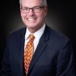 Man in a dark suit with a white shirt and orange patterned tie, smiling against a dark background He has short gray hair and is wearing glasses