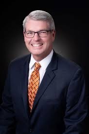 Man in a dark suit with a white shirt and orange patterned tie, smiling against a dark background He has short gray hair and is wearing glasses