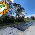 A freshly paved section of road stretches under a clear blue sky, bordered by trees on the left Orange traffic cones are placed on the right A logo in the top left corner displays "Palm Coast Progress