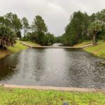 A serene canal with calm water surrounded by lush green grass and pine trees Overcast sky adds a peaceful atmosphere to the scene