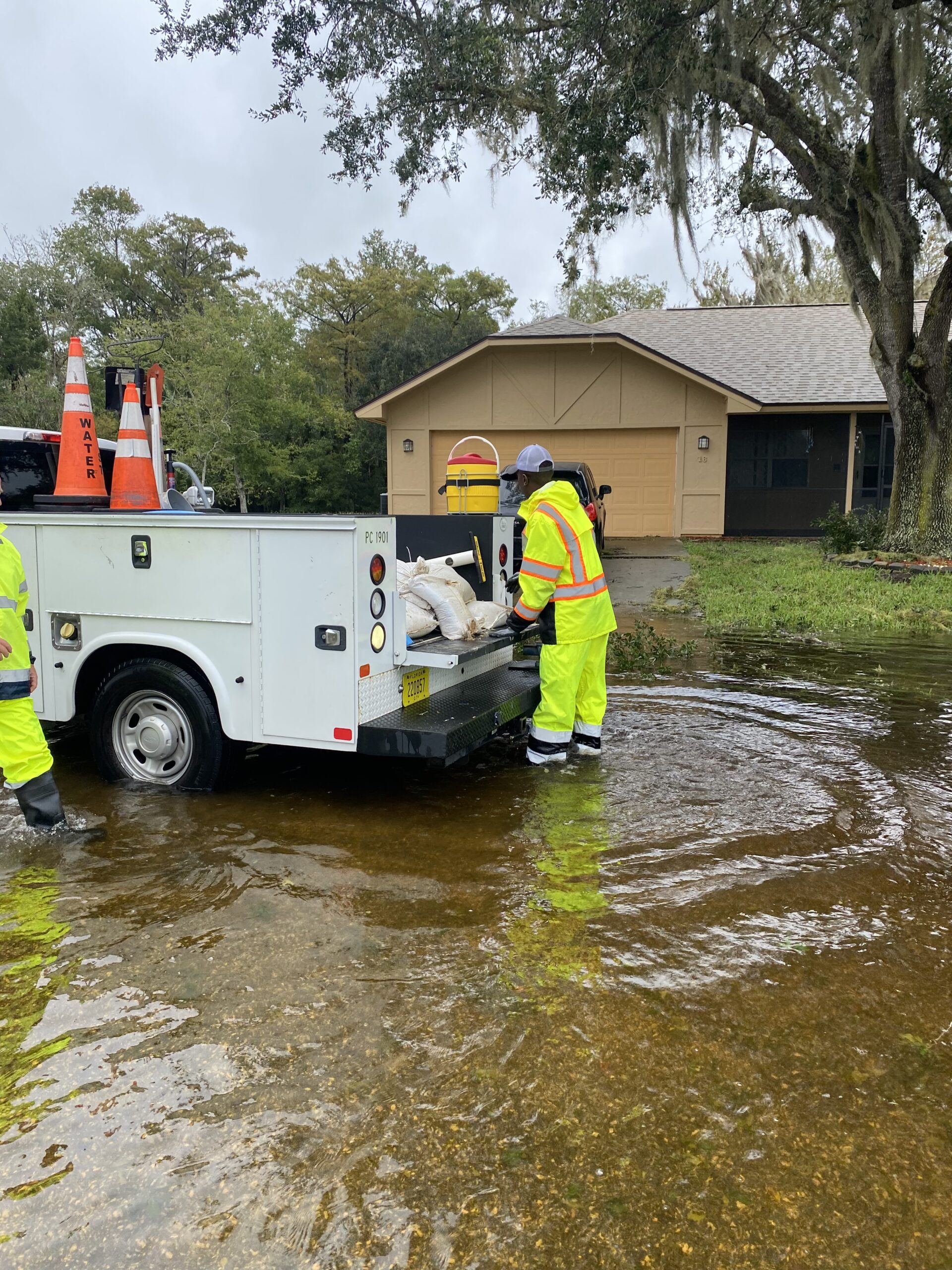 Two workers in neon safety gear are standing in floodwater next to a utility truck, placing sandbags from the truck onto the flooded street The truck has safety cones on it, and a house is visible in the background