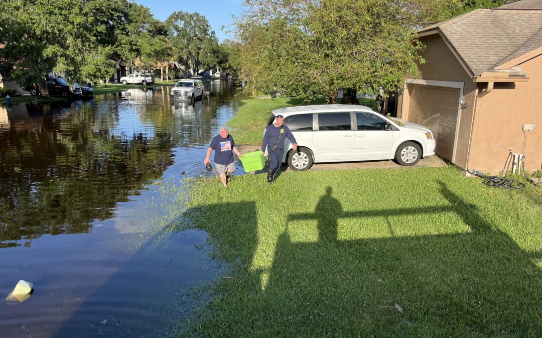 Two men walk through a flooded neighborhood street carrying boxes A white van is parked on a driveway and other vehicles are partially submerged The road is covered in water, and shadows are cast on the grass Trees and houses are in the background