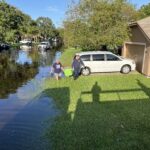 Two men walk through a flooded neighborhood street carrying boxes A white van is parked on a driveway and other vehicles are partially submerged The road is covered in water, and shadows are cast on the grass Trees and houses are in the background