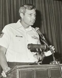 A man in a military uniform speaks passionately at a podium, with microphones in front A name tag and various ribbons are visible on his shirt The setting appears formal, with dark curtains in the background