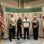 Four people stand in front of a backdrop labeled "Florida Corrections Accreditation Commission," with two flags on either side The man in the center holds a framed certificate Badges and insignias are displayed on the backdrop