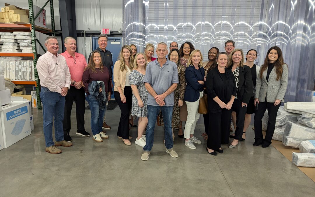 A group of people poses together in a warehouse setting, standing on a concrete floor with shelves and boxes nearby They are dressed in casual and business attire, smiling warmly at the camera