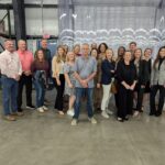 A group of people poses together in a warehouse setting, standing on a concrete floor with shelves and boxes nearby They are dressed in casual and business attire, smiling warmly at the camera