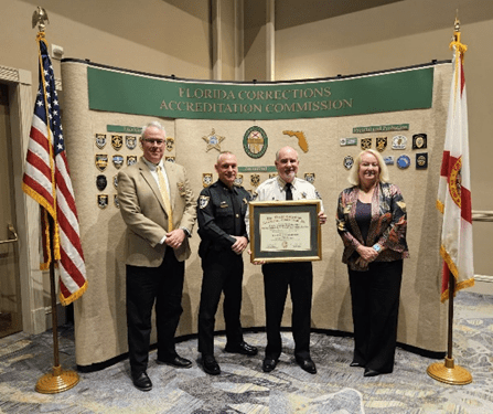 Four people stand in front of a backdrop labeled "Florida Corrections Accreditation Commission," with two flags on either side The man in the center holds a framed certificate Badges and insignias are displayed on the backdrop