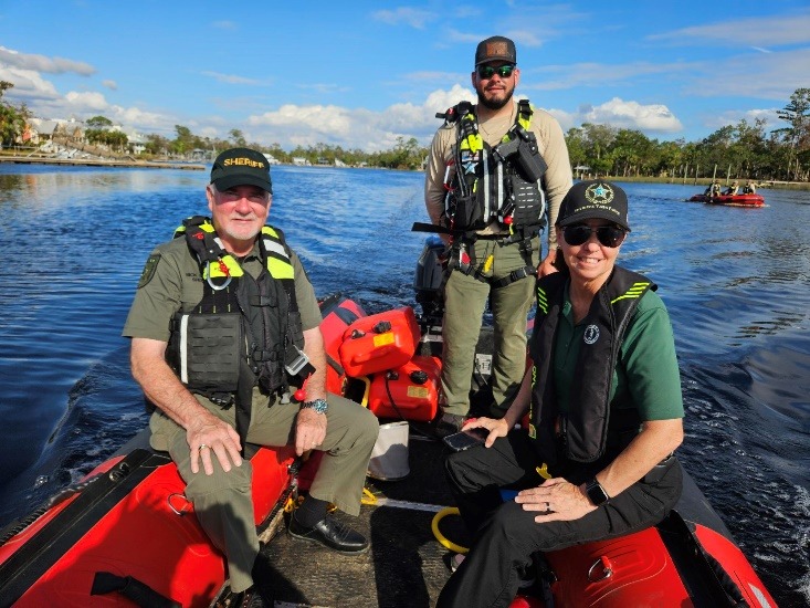 Three people wearing sheriff uniforms and life vests sit and stand on a red inflatable boat on a calm body of water One holds a rescue radio Trees and buildings are visible in the background under a blue sky with clouds