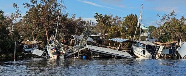 A river scene showing collapsed docks, damaged boats, and debris strewn along the shoreline Trees in the background show signs of damage The sky is partly cloudy