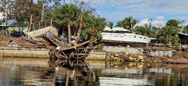 A damaged wooden structure and a large white boat are stranded on a muddy shoreline amidst scattered debris Brown trees and a cloudy blue sky are in the background