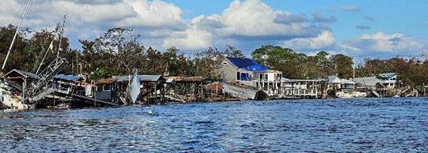 Shoreline with damaged buildings and docks along a body of water Trees are sparse and some have broken branches A few houses have tarps on their roofs The sky is partly cloudy with some sunlight