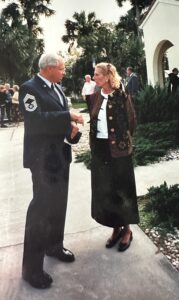 A military officer in uniform converses with a woman wearing a long skirt and jacket outdoors They stand on a pathway surrounded by greenery and palm trees, with a building and other people in the background