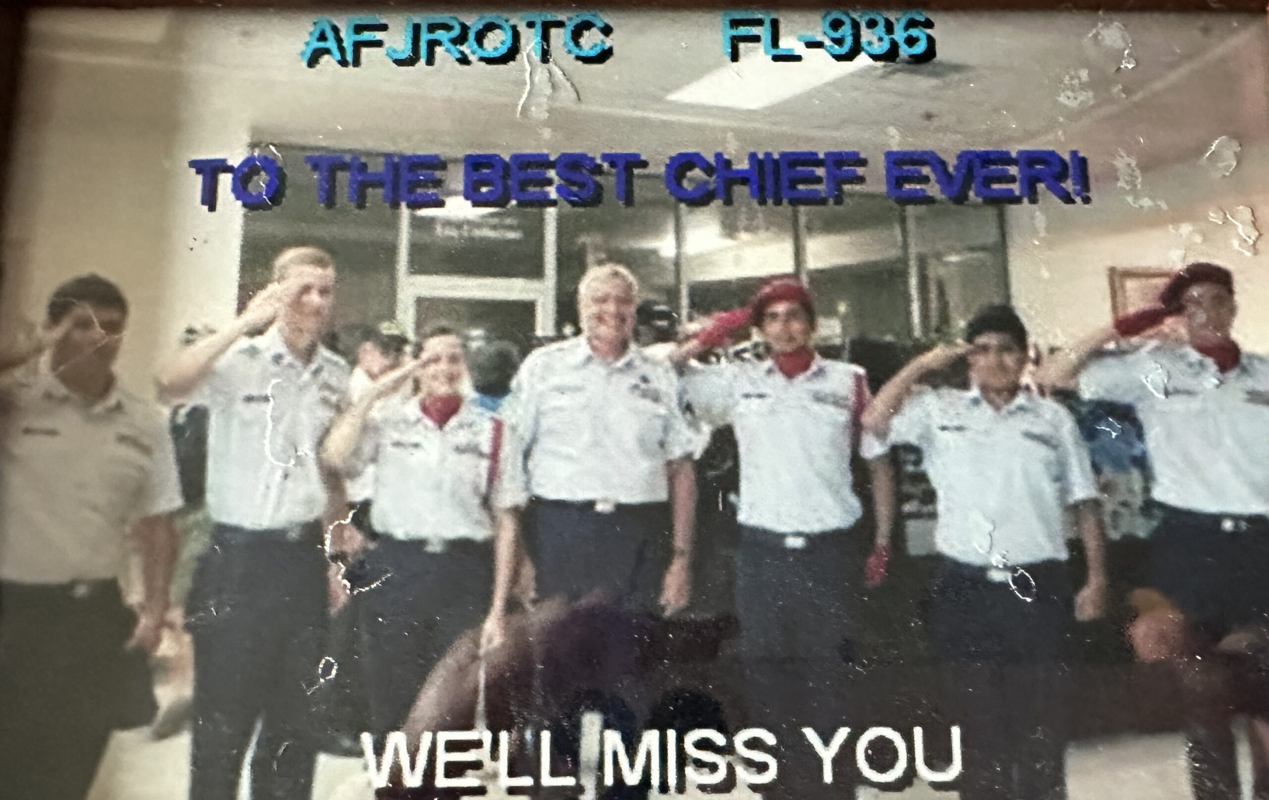 A group of uniformed cadets stands in a line saluting, with the words "AFJROTC FL TO THE BEST CHIEF EVER! WE'LL MISS YOU" overlaid on the image The setting appears to be a room with windows in the background