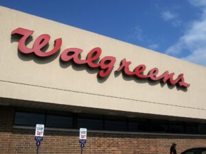 The image shows the exterior of a Walgreens store with its large red logo on a tan background Below are designated parking signs for Walgreens customers A blue sky is visible in the background