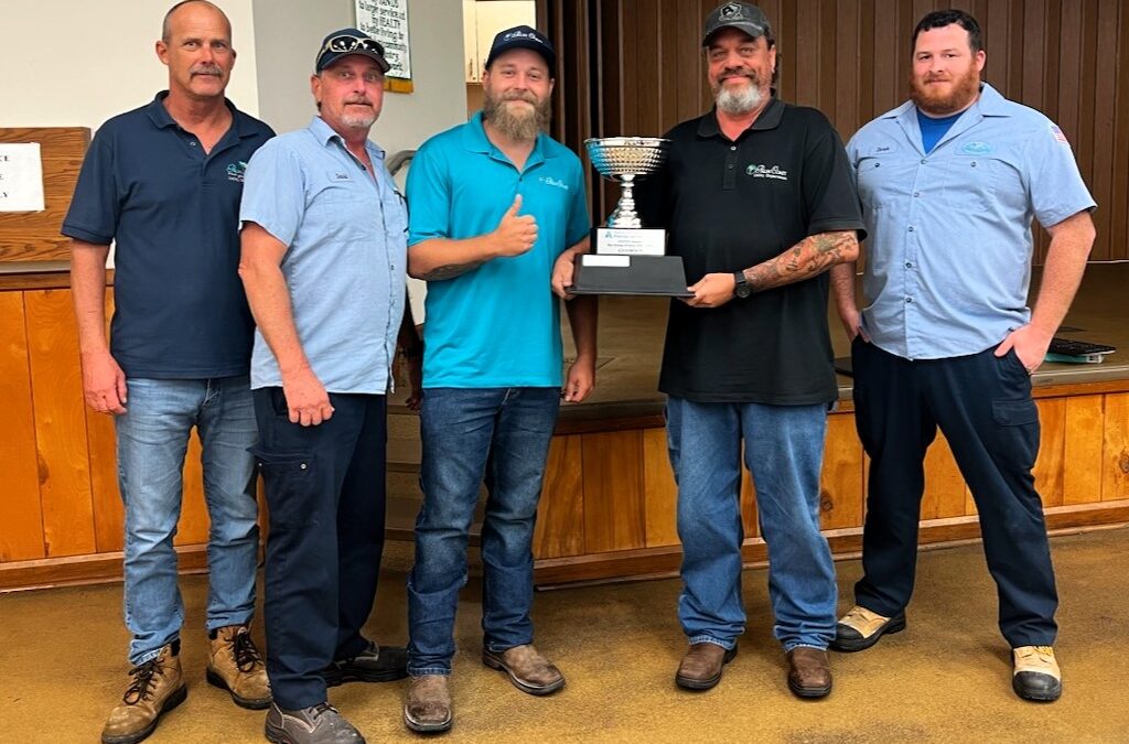 Five men standing in a row indoors, with one holding a trophy They are wearing casual work attire and appear to be on a wooden floor with a stage behind them They are looking at the camera and smiling