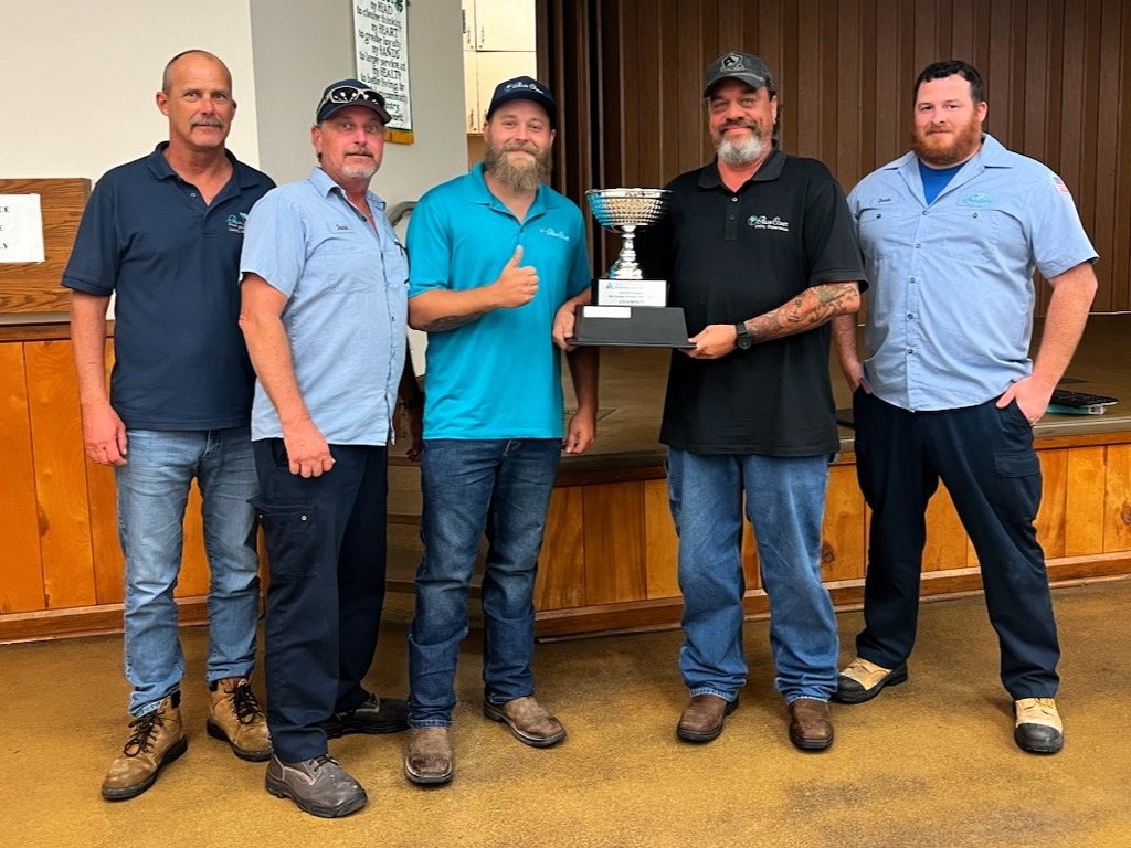 Five men standing in a row indoors, with one holding a trophy They are wearing casual work attire and appear to be on a wooden floor with a stage behind them They are looking at the camera and smiling