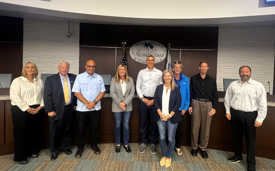 A group of nine people standing in a row, dressed in business and casual attire They are in a room with a curved wooden table and a logo on the wall behind them