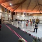 A group of people participating in a dance or exercise class in a spacious hall with a wooden ceiling The instructor stands at the front, leading the participants Various props and personal items are on the floor nearby