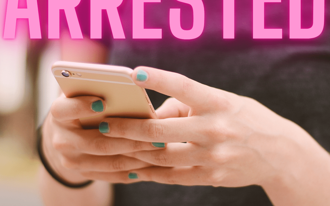 A person holding a smartphone with teal nail polish applied, wearing a dark shirt The word "ARRESTED" is prominently displayed in pink letters at the top of the image