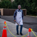 A person wearing a bloody apron and a mask stands in a street near orange traffic cones They hold a large knife prop, and trees are visible in the background, suggesting a horror themed event or costume