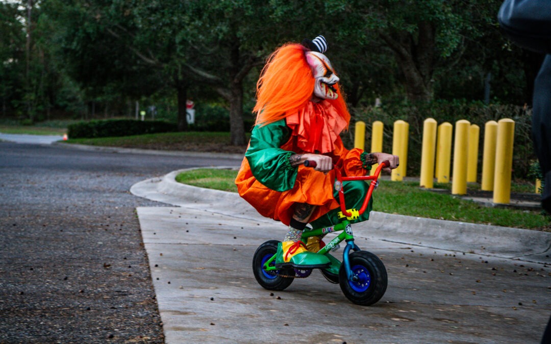 A person dressed as a clown with bright orange hair rides a small green bicycle on a paved path They are in an outdoor setting, surrounded by trees and yellow bollards