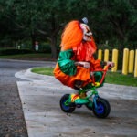 A person dressed as a clown with bright orange hair rides a small green bicycle on a paved path They are in an outdoor setting, surrounded by trees and yellow bollards