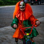 A person in a colorful, spooky clown costume with bright orange hair and a creepy mask rides a small bicycle on a paved path The clown wears a green and orange outfit with exaggerated features, set against a background of trees and a parked truck