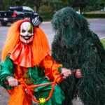 A person in a colorful clown costume with an orange wig and mask rides a small bike Beside them, another person wears a ghillie suit, blending into the background foliage They are on a paved residential street with trees and parked cars