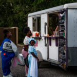 A woman and two children stand by a food truck The woman carries a child while the other child holds a large inflatable costume The food truck menu is visible, and trees are in the background