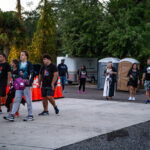 A group of people walking outdoors on a paved path surrounded by trees They are casually dressed, and some are carrying backpacks Orange traffic cones are visible on the left side, and portable toilets are seen in the background