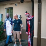 A person in a t shirt and shorts stands outside a building, flanked by two individuals in creepy costumes One wears a blood stained apron and mask, and the other is dressed in a blood splattered clown outfit