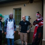 Four people stand outside a building, three dressed in horror themed costumes with bloody aprons and masks One person wears a blood stained clown outfit Another person in casual clothing looks on They appear to be engaged in conversation