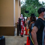 A person in a blood stained costume and mask walks past a line of people outside a building Orange cones and yellow rope mark the path Other people in line wear casual clothes and hats Trees are visible in the background