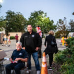 A man sits on a chair at a table, while another person wearing a black jacket stands nearby, smiling Orange traffic cones are on the path Several people are walking in the background, and trees are visible under a bright blue sky