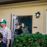 A group of people, including children and adults, are exiting a building through a door They are outdoors with greenery around them, under a beige building with a window and a wall lamp Casual attire suggests a relaxed setting