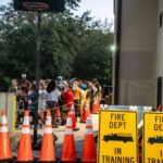 A group of people stand in line outside a building Orange cones and two signs reading "Fire Dept in Training" are visible Trees are in the background, and a basketball hoop is on the left It appears to be early evening with dim lighting