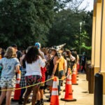 A crowd of people, some wearing costumes, stand in line outside a building bordered by orange traffic cones The scene is set near dusk, with trees in the background and outdoor lights providing illumination