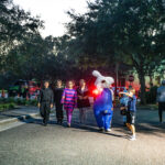 A group of children in Halloween costumes walk along a street in the evening They are surrounded by trees and dim lighting, with one child dressed as the Pillsbury Doughboy holding a red light