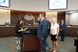 A group of people standing in a room with "Palm Coast" signage in the background One person is being sworn in by a woman holding a document, while others watch The room has a curved desk and flags on display